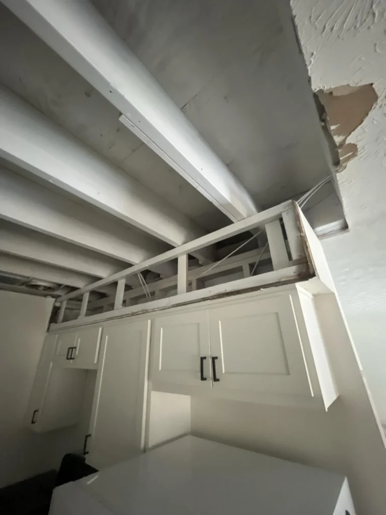 Upward view in a kitchen showing upper white cabinets with black hardware, soffit area above with drywall removed revealing painted wood framing, and a section of drywall with peeling paint suggesting water damage