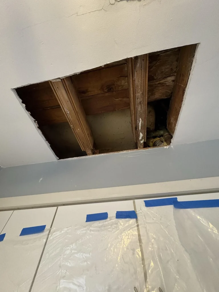 Rectangular drywall cutout in a kitchen ceiling exposing dark water-stained wooden joists, subfloor boards, and visible plumbing pipes, with blue painter's tape and plastic sheeting protecting the white cabinets below
