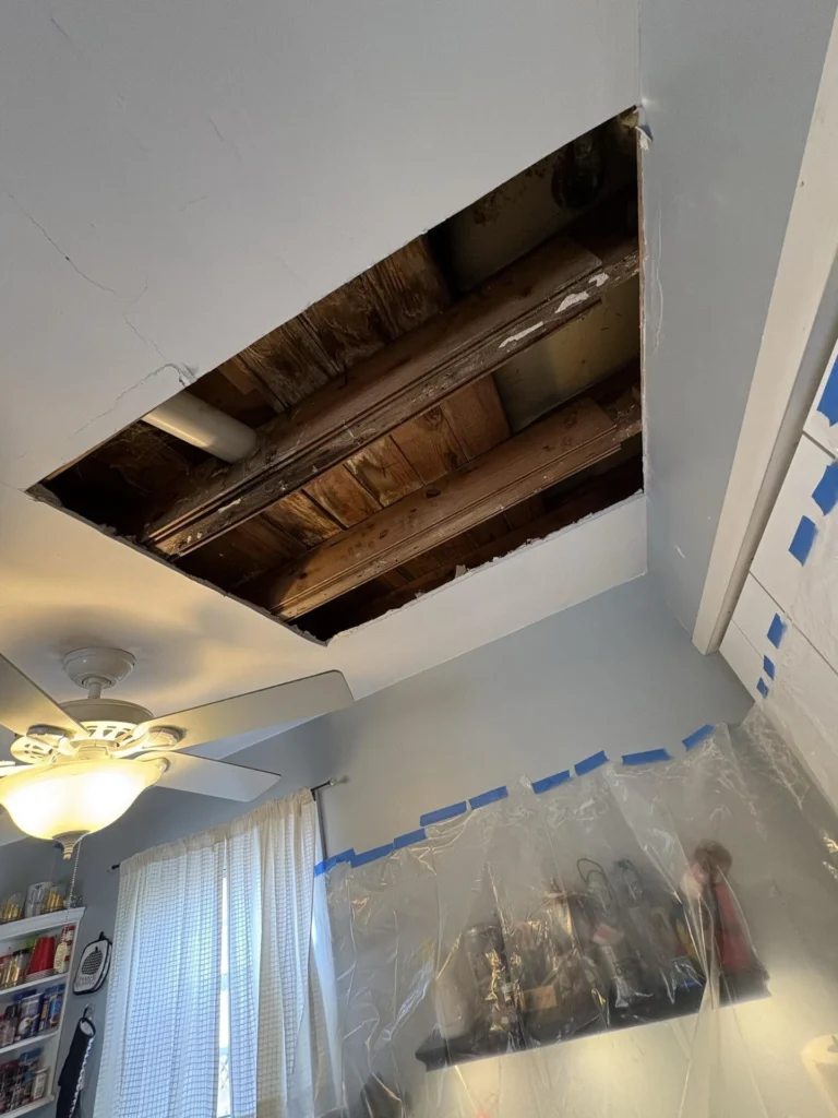 Kitchen ceiling with a large rectangular section of drywall removed exposing dark water-stained wooden joists and subfloor above, with blue painter's tape securing plastic sheeting over cabinetry and a ceiling fan with light visible