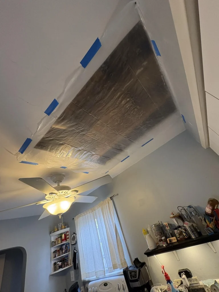Close-up of a kitchen ceiling with a large silver foil-faced water containment barrier secured with blue painter's tape around the perimeter, above a ceiling fan and light fixture, with shelving and countertop visible below