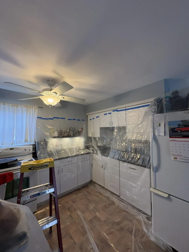 Kitchen showing ceiling water damage with cabinets and countertops wrapped in clear plastic sheeting held with blue tape, a ceiling fan and light, a white refrigerator, and a Werner ladder visible during water damage restoration work