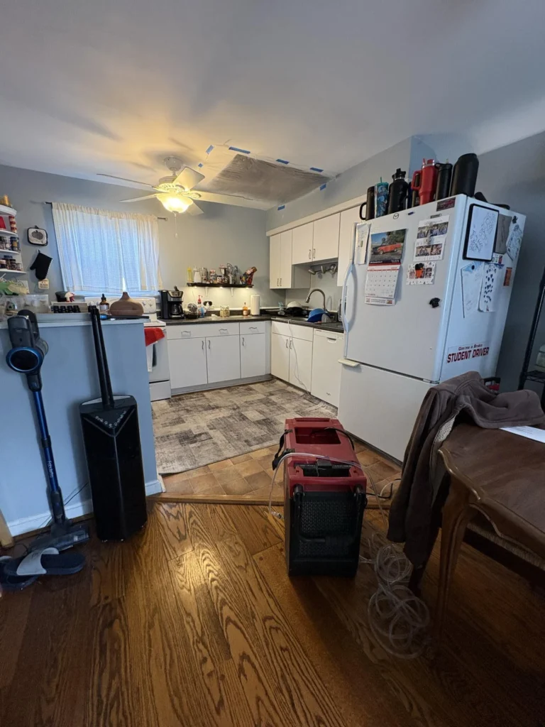 Kitchen with ceiling water damage showing a large area covered with silver foil-faced barrier taped with blue painter's tape above a ceiling fan, with a red dehumidifier on the floor and white cabinetry and refrigerator visible