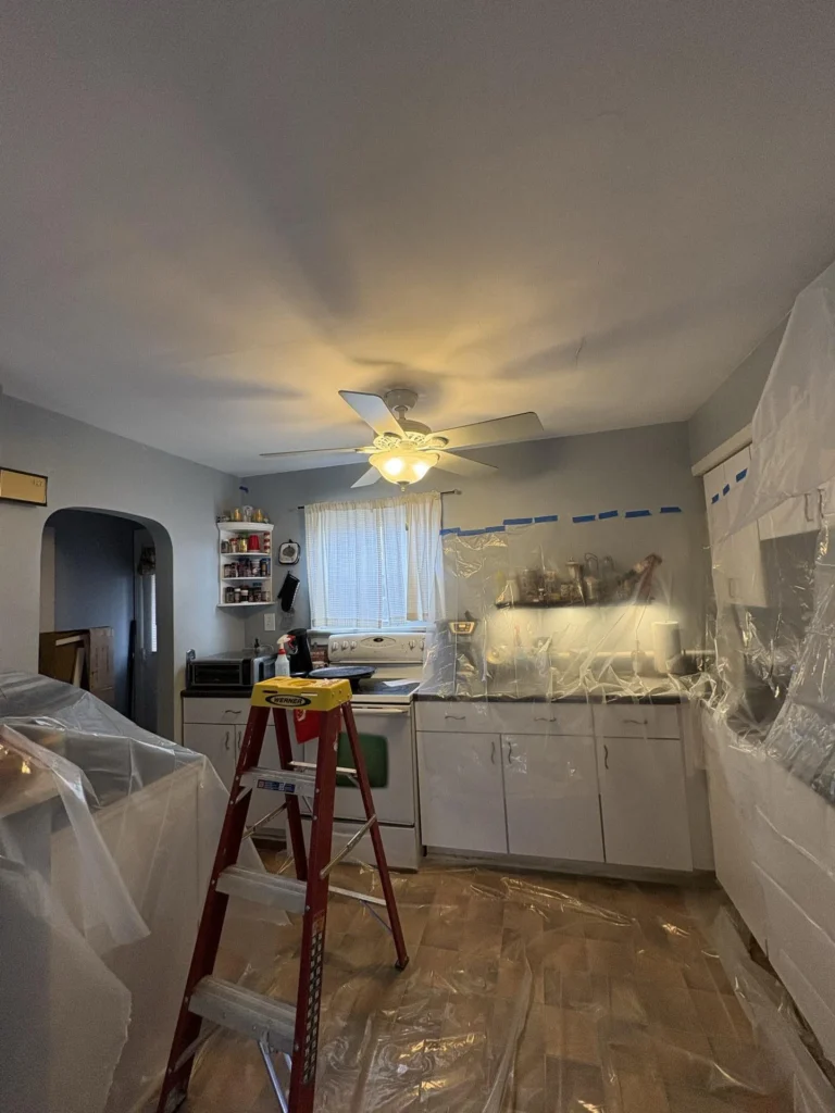 Kitchen during water damage remediation with white cabinets and countertops covered in plastic sheeting secured with blue tape, a red Werner step ladder in the center, and water stain discoloration visible on the ceiling