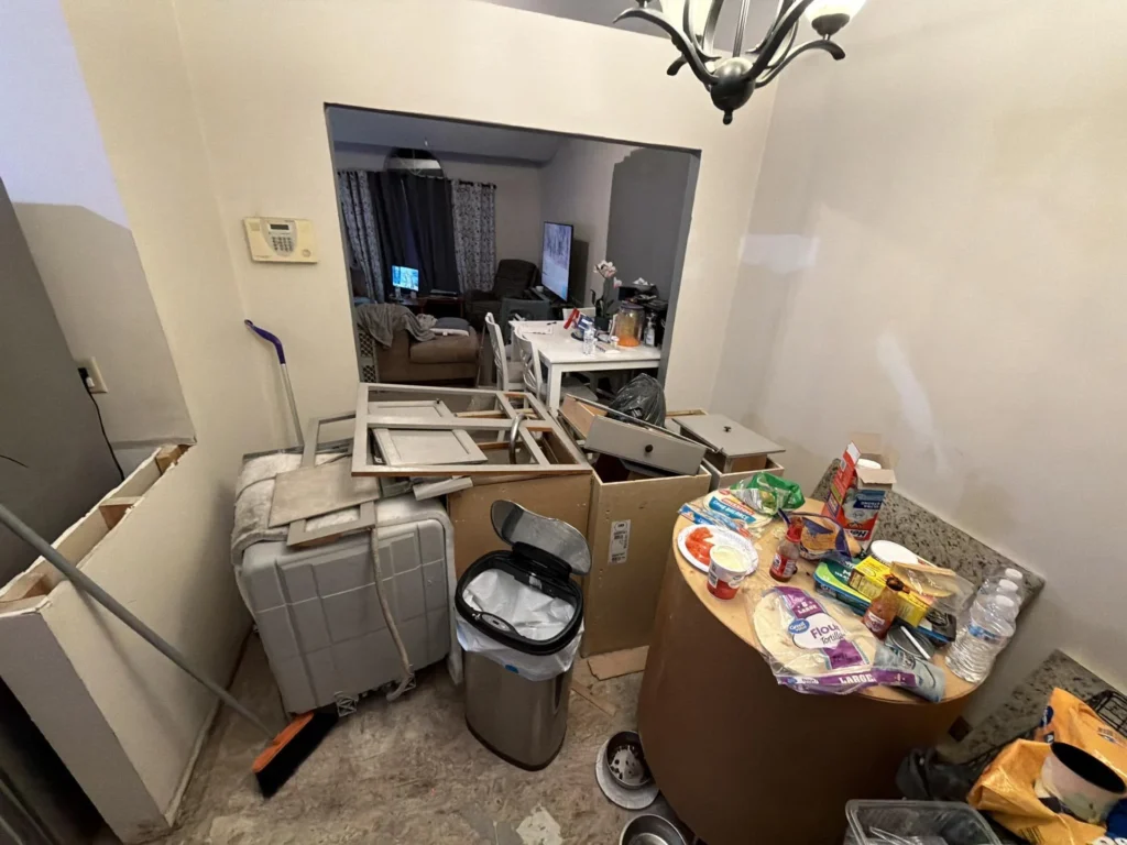 Flood-damaged home interior with demolished kitchen cabinets and doors piled in the dining area, suitcase and trash can amid debris, food items on a round table, open pass-through to living room with TV visible