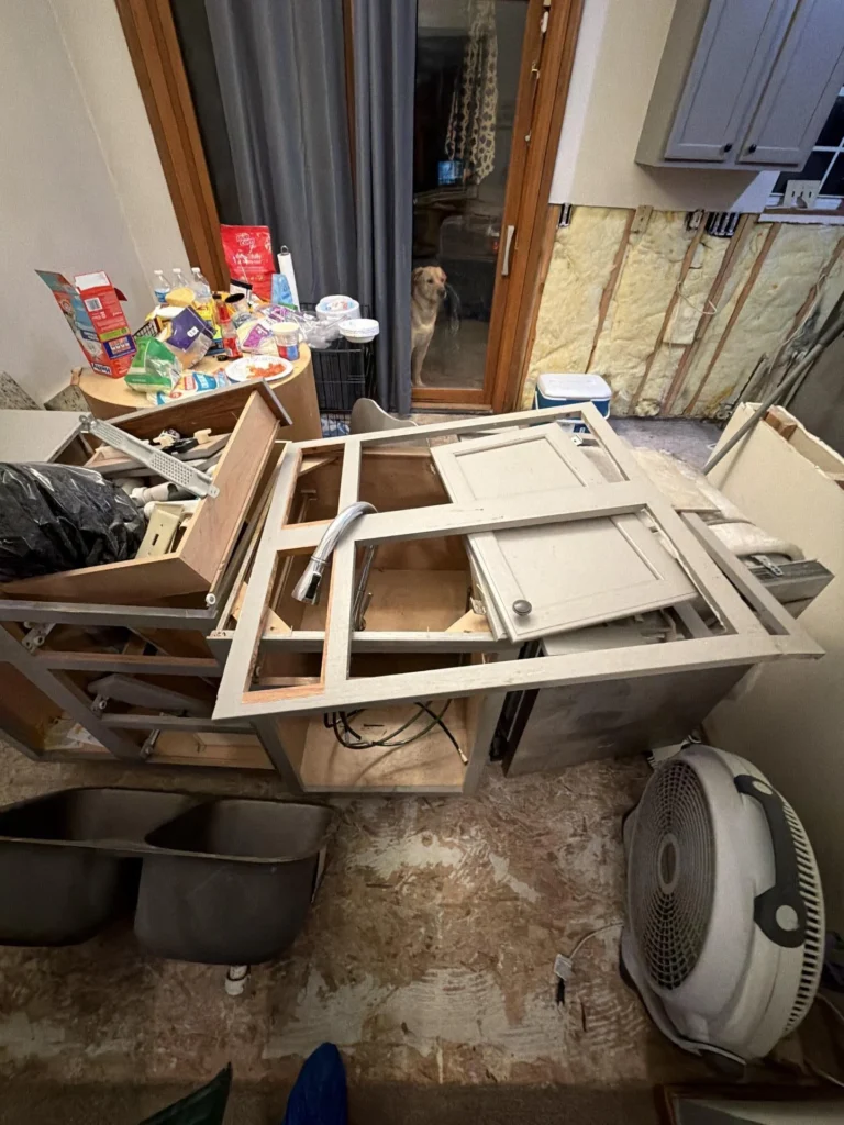 Overhead view of a flood-damaged kitchen with base cabinets removed and tipped over, exposed subfloor, detached sink, cabinet doors and hardware scattered, exposed wall insulation, and a box fan on the floor