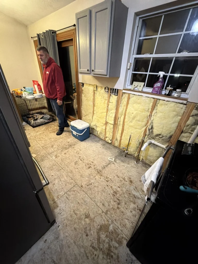 Water-damaged kitchen with lower wall drywall removed exposing yellow fiberglass insulation and plumbing pipes, worker in red hoodie standing in doorway, stove with food cooking, dog crate visible in background