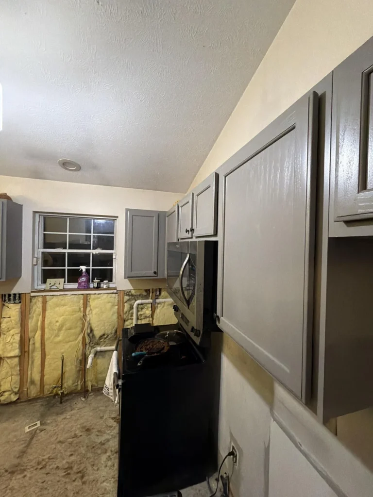 Flood-damaged kitchen viewed from below showing gray upper cabinet doors, stainless over-range microwave, black electric stove with pan of food, and fully exposed yellow fiberglass insulation on lower walls