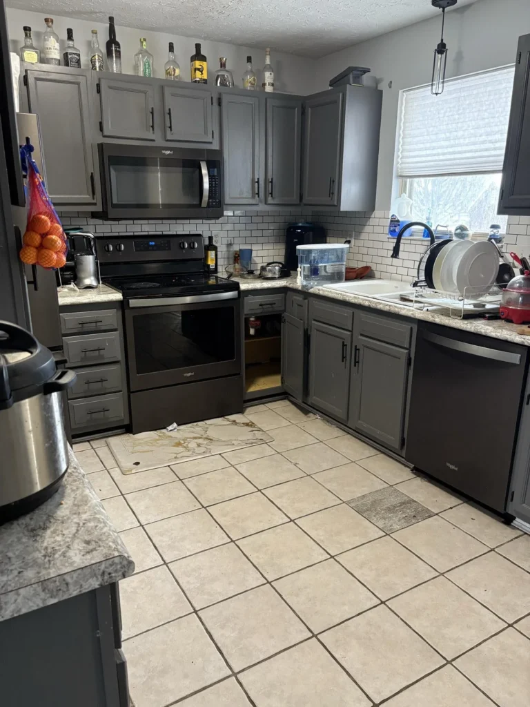 Full kitchen view showing gray cabinets, Whirlpool black stainless range with over-range microwave, stainless dishwasher, white subway backsplash, and water staining on the tile floor below the range