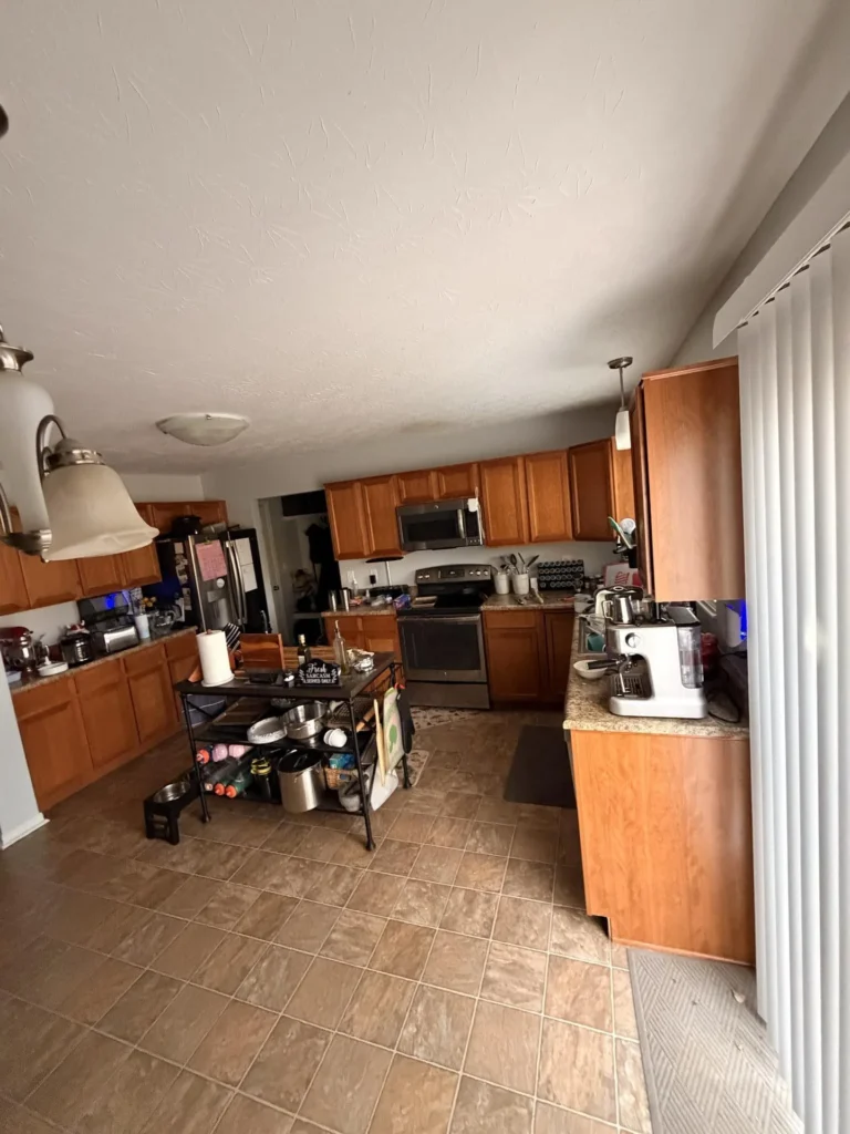 Spacious kitchen with oak wood cabinets, granite countertops, tile floor, stainless steel appliances including range with microwave above, refrigerator, and pendant lighting, with a black wire shelving cart as a portable island
