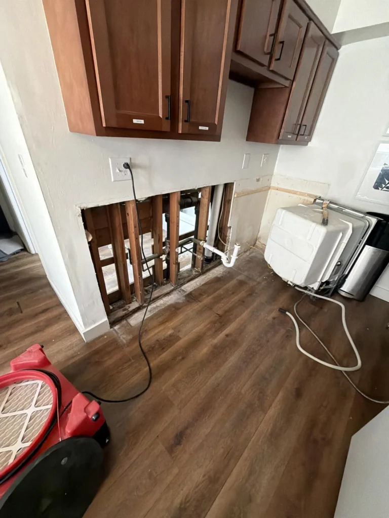 Kitchen showing dark wood upper cabinets with lower wall section removed exposing multiple PVC and metal drain pipes inside wall cavity, red air mover and white appliances on hardwood floor during water damage drying