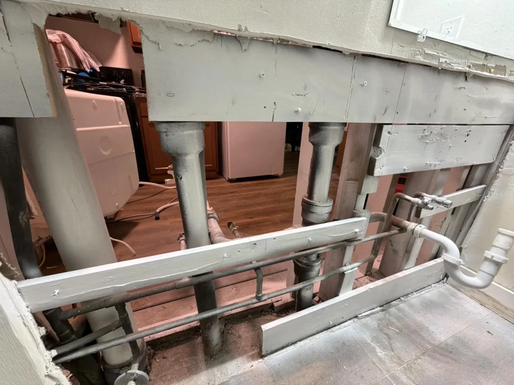 Large rectangular opening cut in a wall between bathroom and kitchen exposing cast-iron drain pipes, galvanized water supply lines, and wood framing, with kitchen appliances and hardwood flooring visible through the opening