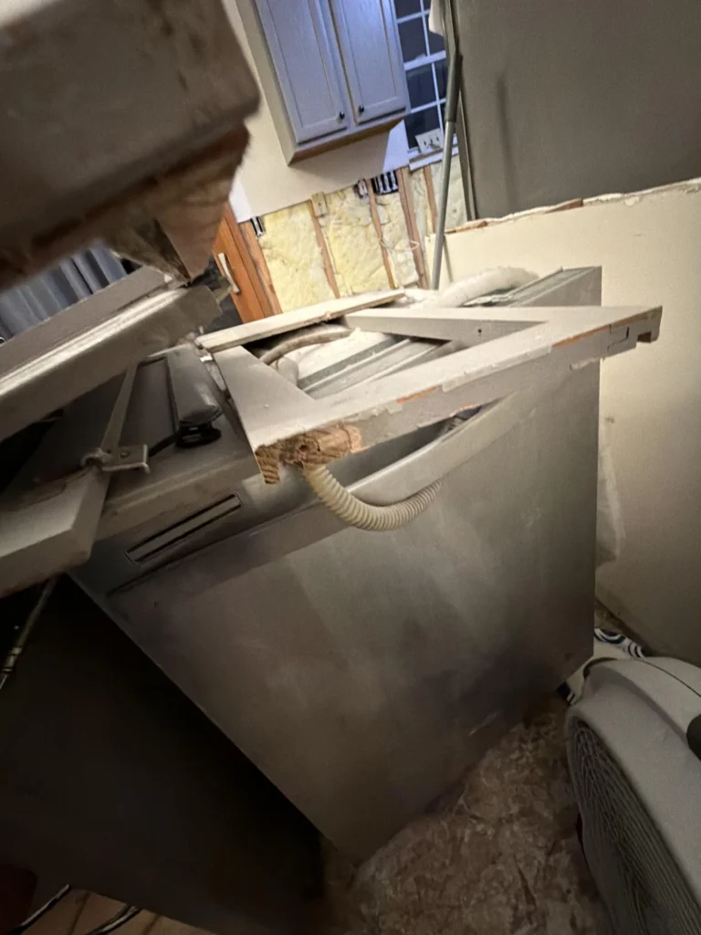 Close-up of a water-damaged kitchen showing a dishwasher pulled out with drain hose exposed, demolished base cabinet frames piled up, exposed fiberglass wall insulation, and damaged subfloor