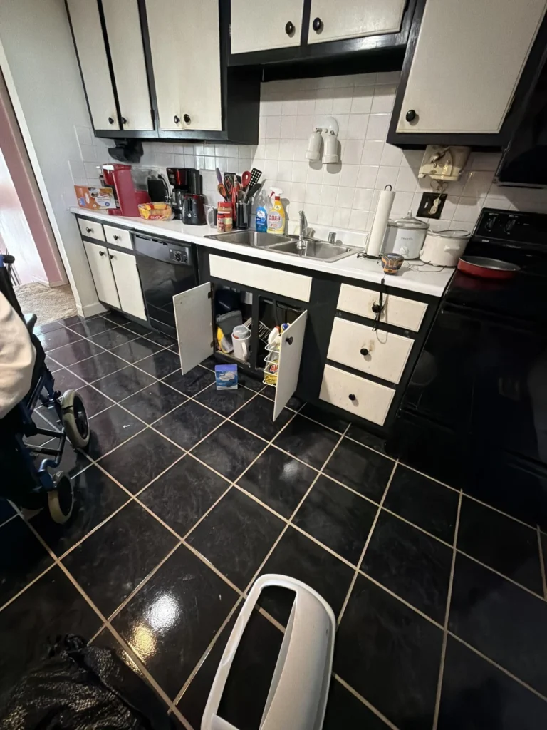 Kitchen with black and white cabinetry showing water damage with the under-sink cabinet door open revealing wet contents, black tile floor, countertop with appliances and cleaning supplies, and a wheelchair visible to the left