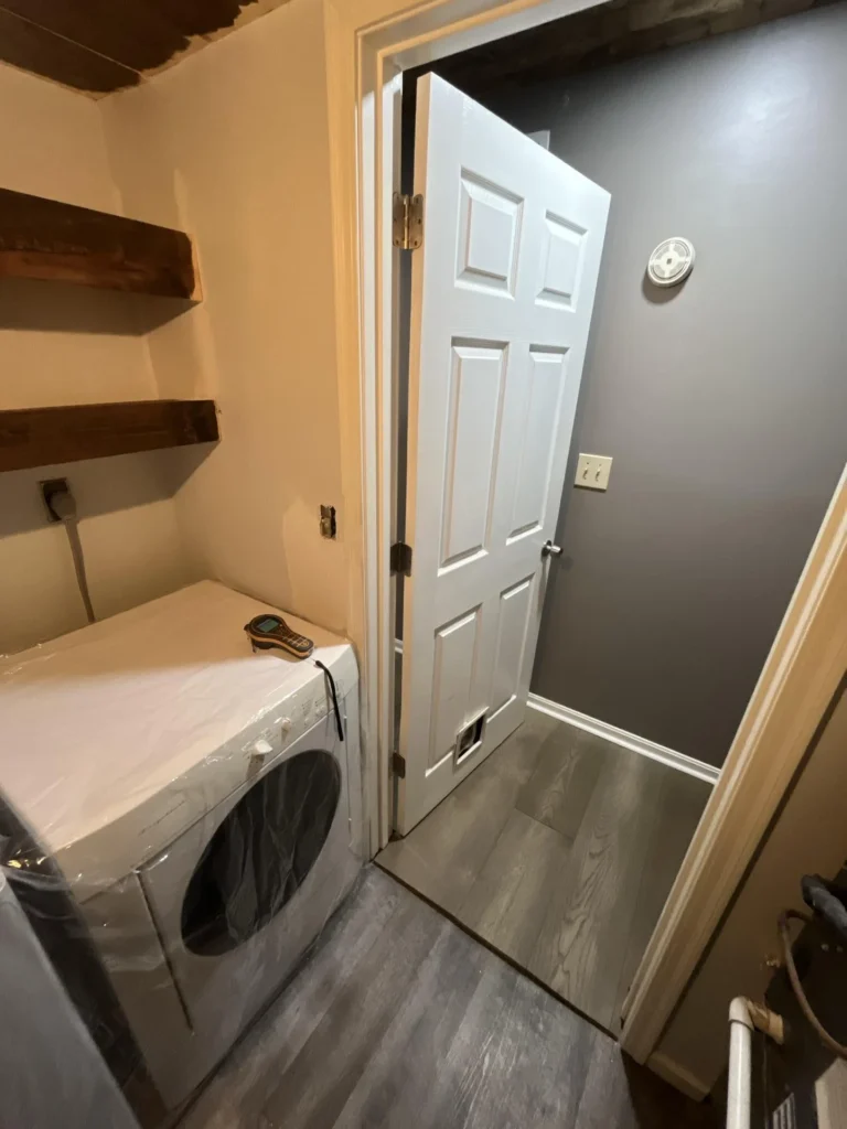 Laundry room interior showing a front-load dryer in plastic wrap, floating wood shelves, open door leading to a gray hallway with LVP flooring, and a moisture meter on top of the dryer