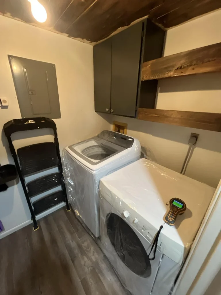 Full view of a laundry room with top-load washer and front-load dryer wrapped in plastic, dark gray upper cabinets, rustic wood floating shelves, electrical panel on wall, and step stool