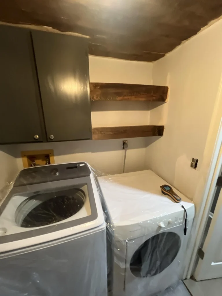 Laundry room with a new top-load washer and front-load dryer still in plastic wrap, dark gray upper cabinets, floating wood shelves, and a water-damaged wood plank ceiling above