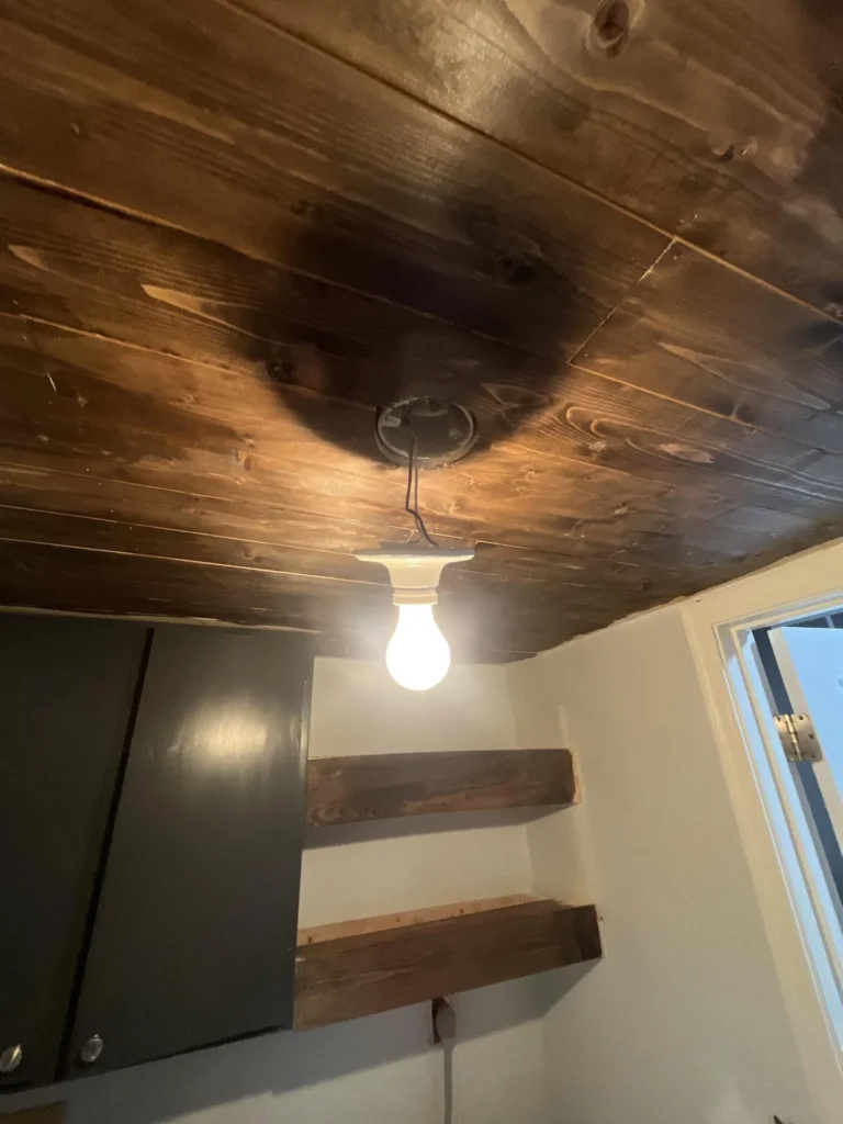 Laundry room ceiling showing dark water staining on wood plank boards above a bare bulb light fixture, with floating wood shelves and gray cabinets visible below the damage