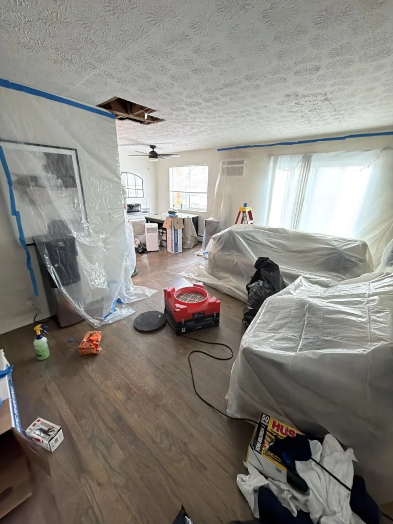 Living room during water damage remediation with furniture covered in plastic sheeting, a square ceiling opening exposing joists, red air scrubber on the floor, blue-taped plastic containment barriers, and a stepladder