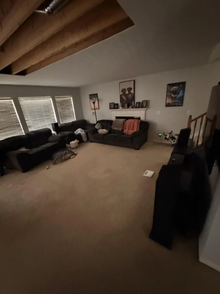 Living room with dark sectional sofa and couch around a white fireplace with framed artwork above, beige carpet, large windows with blinds, and significant water damage visible on the ceiling with large section of drywall removed exposing wooden joists
