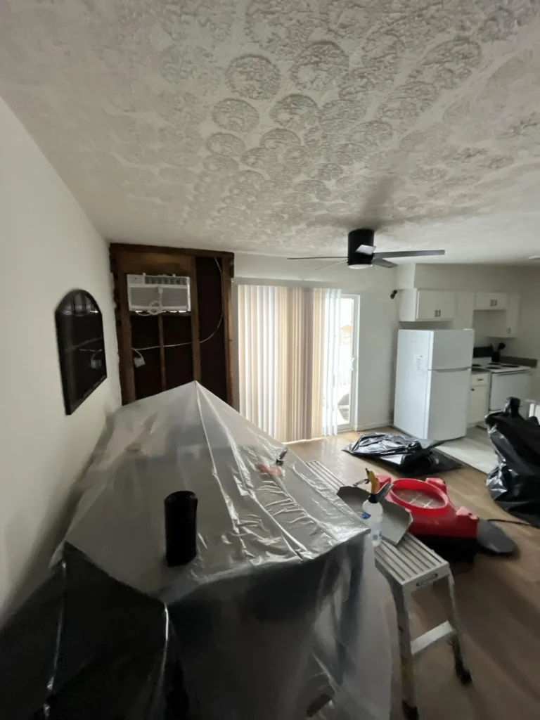 Living room during mold remediation with furniture covered in plastic sheeting, large section of exterior wall drywall removed exposing dark wood framing and window unit AC, popcorn ceiling with staining, red air scrubber and containment barriers