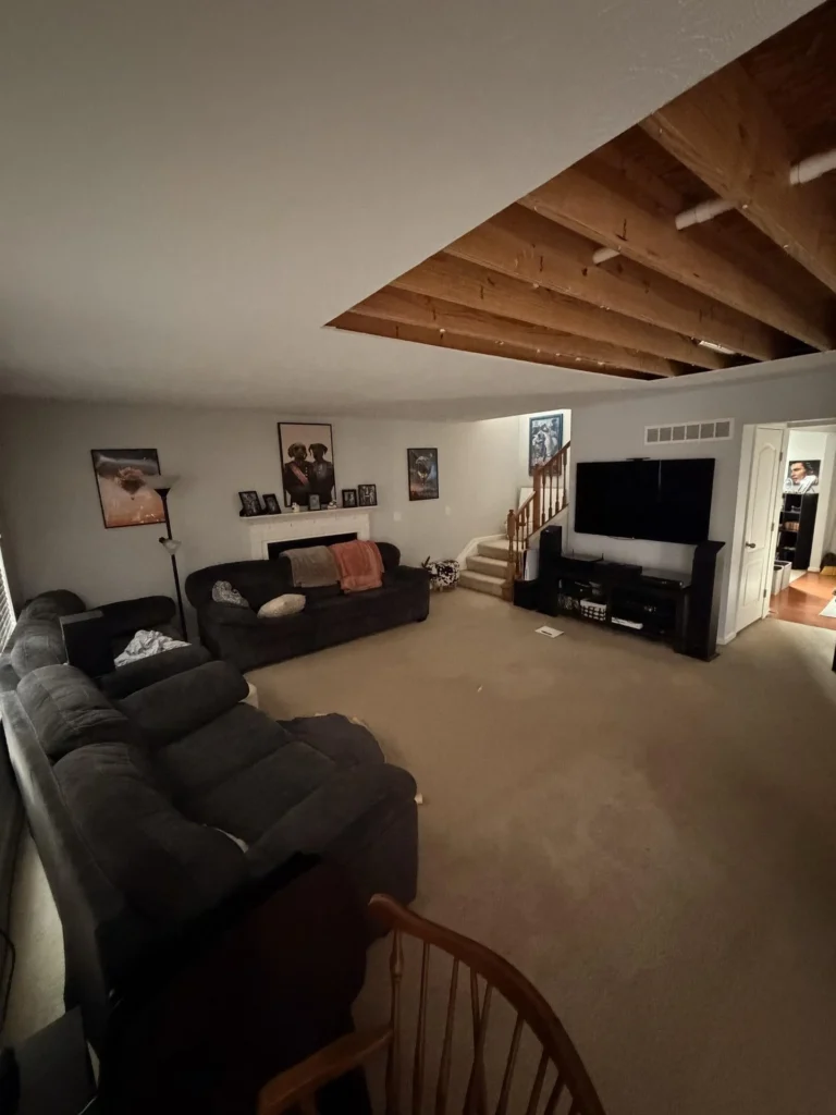 Large living room with dark sectional sofa, large flat-screen TV and entertainment center, white fireplace with framed art, beige carpet, staircase with oak railing, and large section of ceiling drywall removed exposing wood joists due to water damage