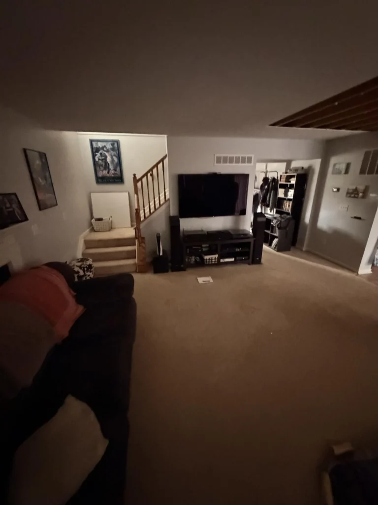 Lower level living room with dark sectional sofa, large flat-screen TV on entertainment center with speakers, wood stair railing leading upstairs, framed posters, shelving unit with books and media, beige carpet, and open doorway to adjacent room