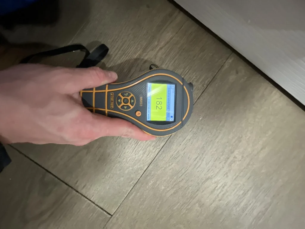 Technician holding a pinless moisture meter displaying an elevated reading of 182 on a yellow-green screen while testing luxury vinyl plank flooring in a residential hallway