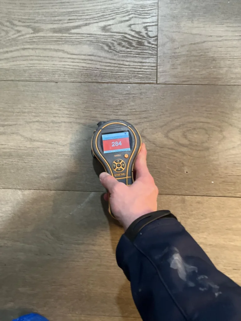 Technician holding a pinless moisture meter displaying an elevated reading of 284 on a red screen while testing luxury vinyl plank flooring during a water damage inspection