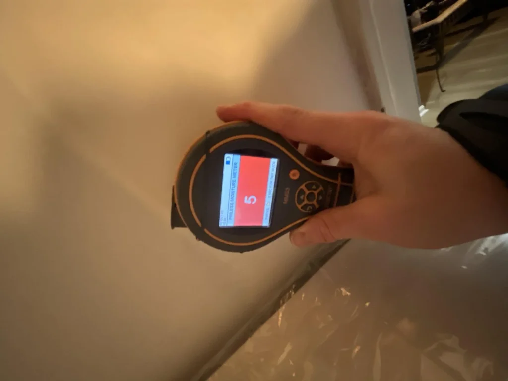 Technician holding a pinless moisture meter displaying a low reading of 5 on a red screen while testing a painted ceiling surface near a plastic vapor barrier during water damage remediation