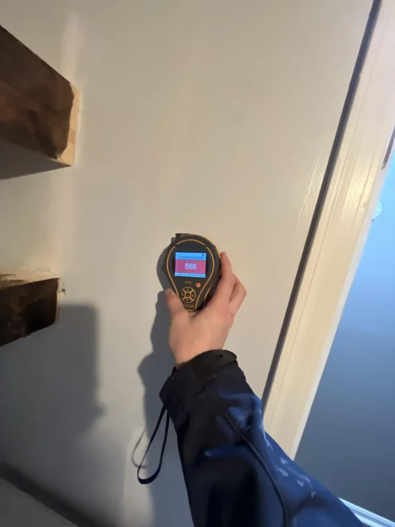Technician holding a pinless moisture meter showing an elevated reading of 568 pressed against a white drywall surface near a door frame during a water damage inspection