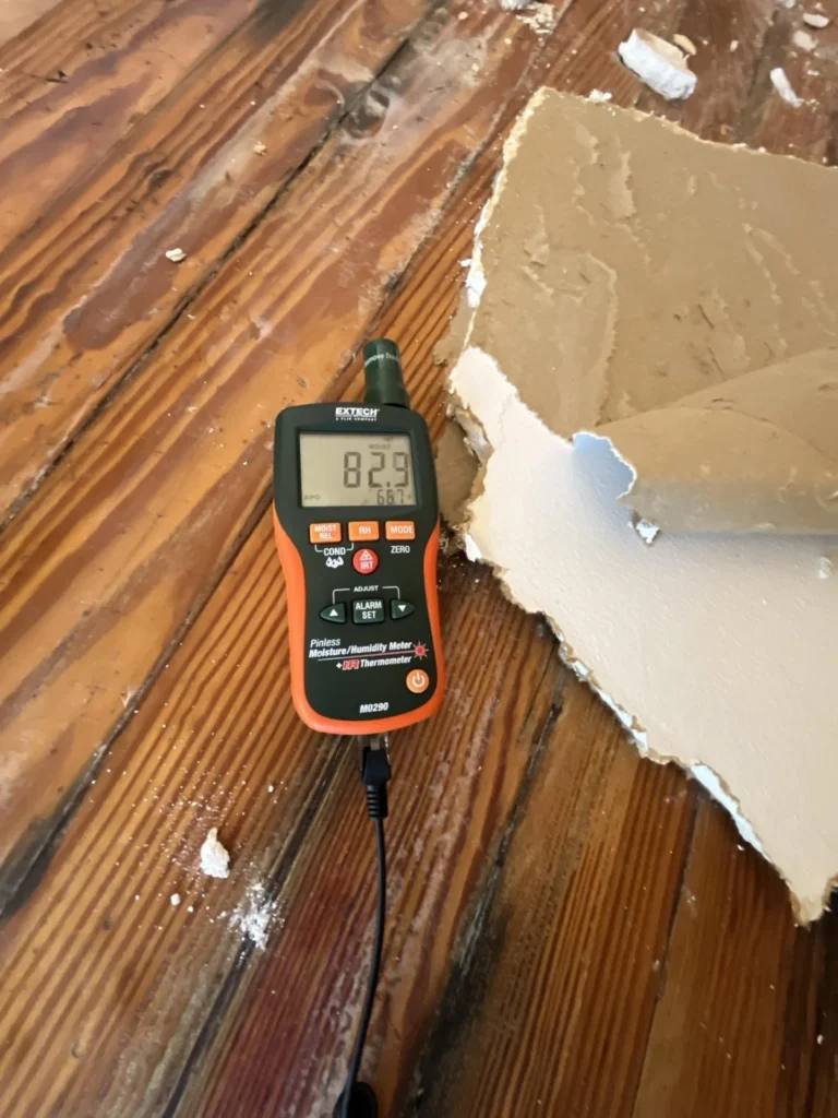 An Extech M0290 pinless moisture and humidity meter displaying a reading of 82.9 percent moisture, placed on water-damaged hardwood floorboards next to a fallen piece of drywall