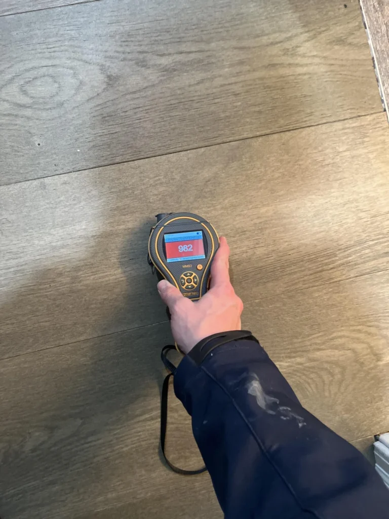 Technician holding a pinless moisture meter displaying a high reading of 982 on a red screen while testing water-damaged luxury vinyl plank flooring during a water damage assessment
