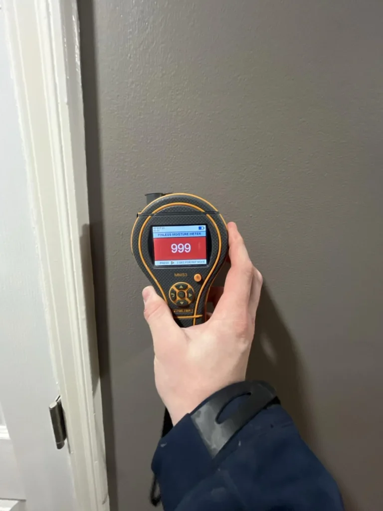 Close-up of a pinless moisture meter showing a maxed-out reading of 999 held against a gray drywall surface next to a white door frame during a water damage assessment