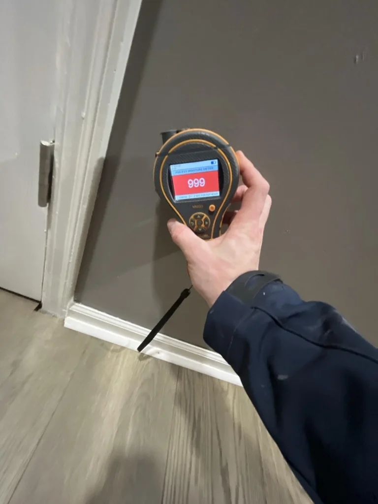 Technician holding a pinless moisture meter displaying a maximum reading of 999 against a gray wall at the baseboard level during a water damage inspection in a residential hallway