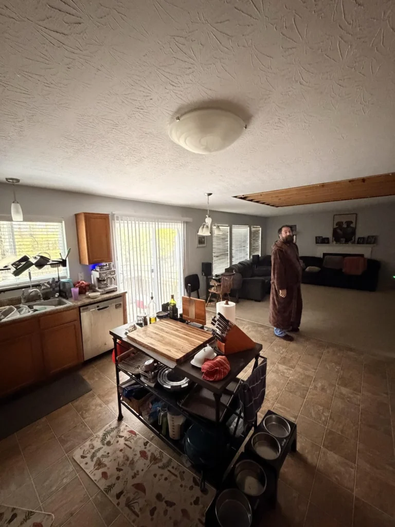 Open-plan kitchen, dining, and living room with person in robe looking at large section of ceiling showing water damage with exposed wood joists, oak cabinets, sink, dishwasher, sliding patio door, rolling cart, and dark furniture beyond