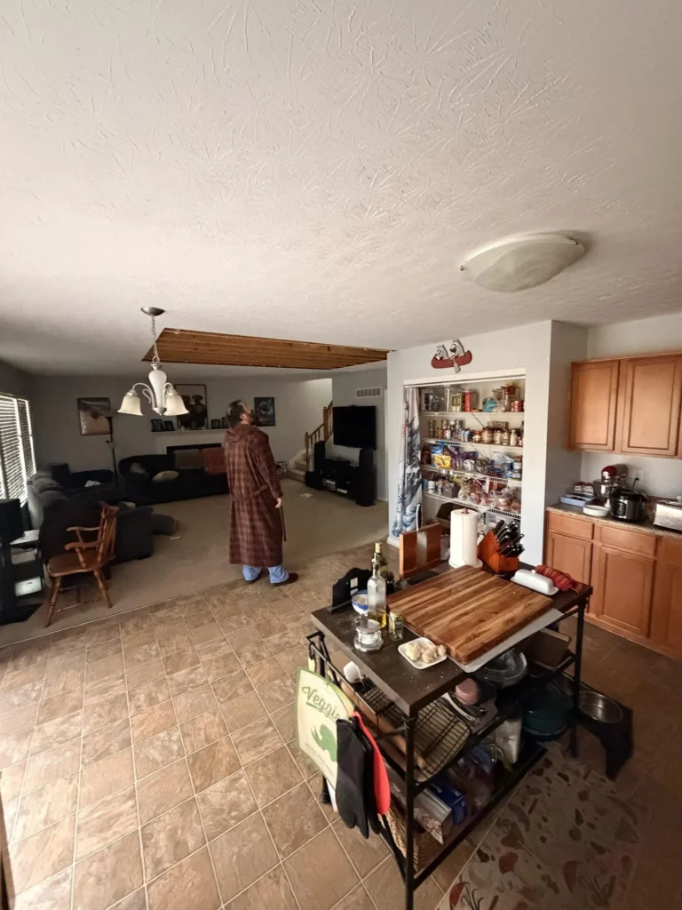 Open-plan kitchen and living area with person in robe looking at ceiling water damage where drywall is removed showing wood structure, oak cabinets visible, rolling cart, wall shelving with pantry items, and living room with TV and sofa beyond