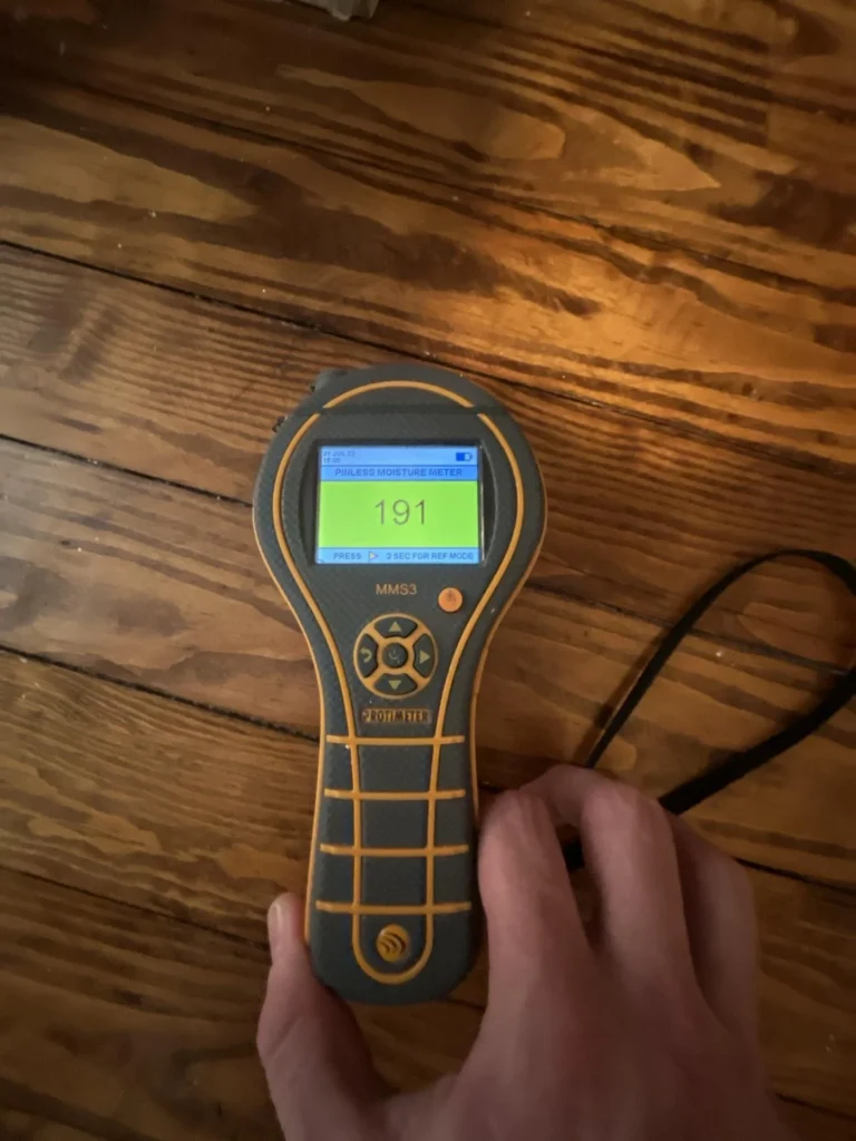 Hand holding a Protimeter MMS3 pinless moisture meter displaying a reading of 191 on a green screen, pressed against wood subfloor planks seen from below