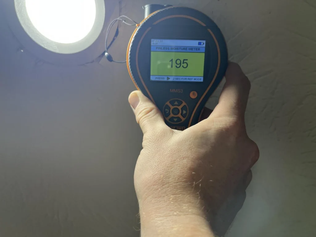 Hand holding a Protimeter MMS3 pinless moisture meter displaying a reading of 195 on a green screen, pressed against a textured white ceiling near a recessed light fixture