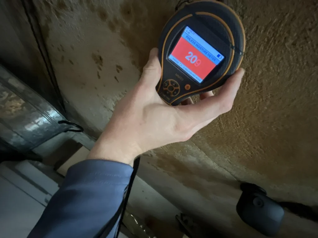 Hand holding a Protimeter MMS3 pinless moisture meter displaying a reading of 209 on a red screen, pressed against a heavily water-stained and discolored garage ceiling
