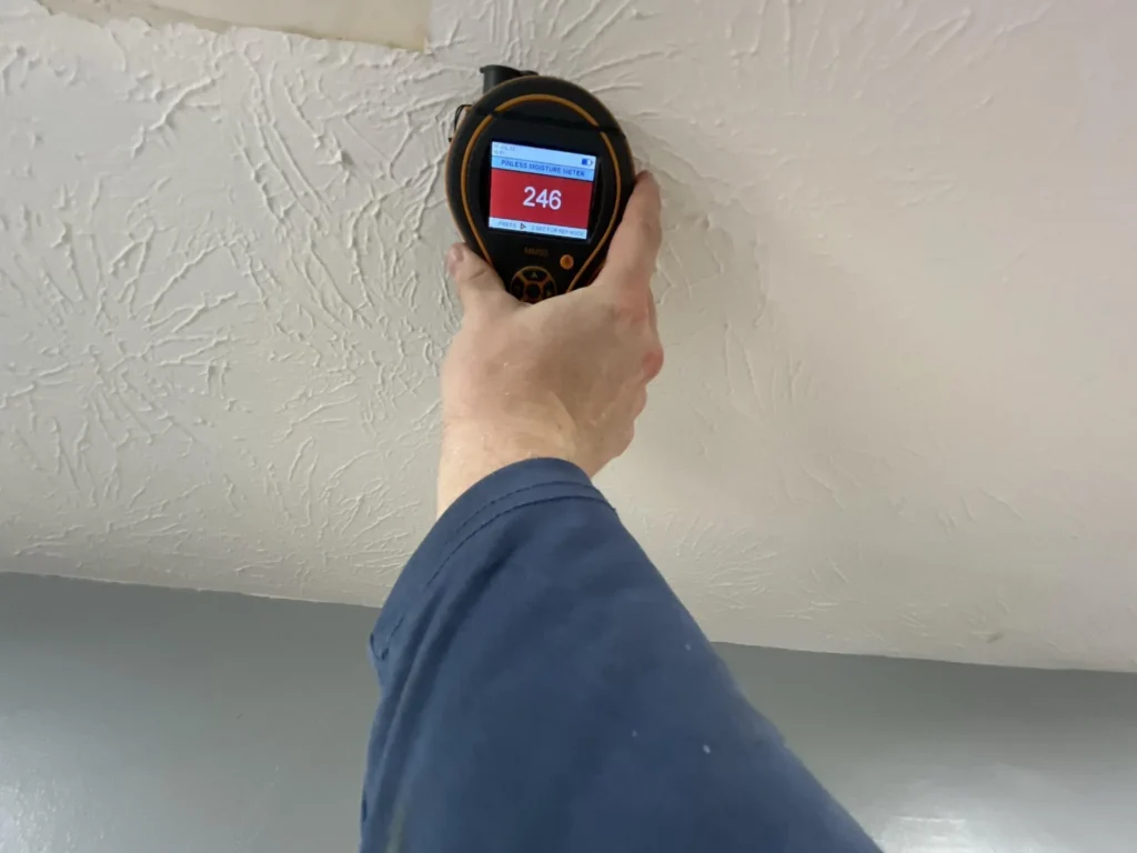 Hand and arm holding a Protimeter MMS3 pinless moisture meter displaying a reading of 246 on a red screen, pressed against a textured white ceiling