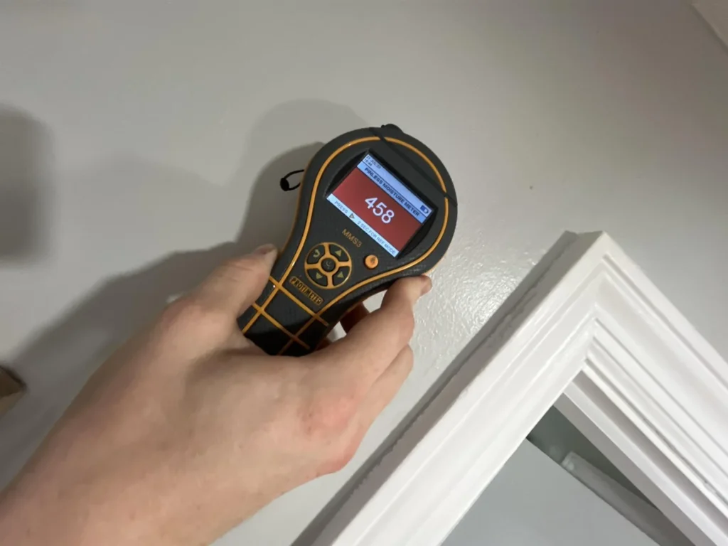 Hand holding a Protimeter MMS3 pinless moisture meter displaying a reading of 458 on a red screen, pressed against a white interior door frame