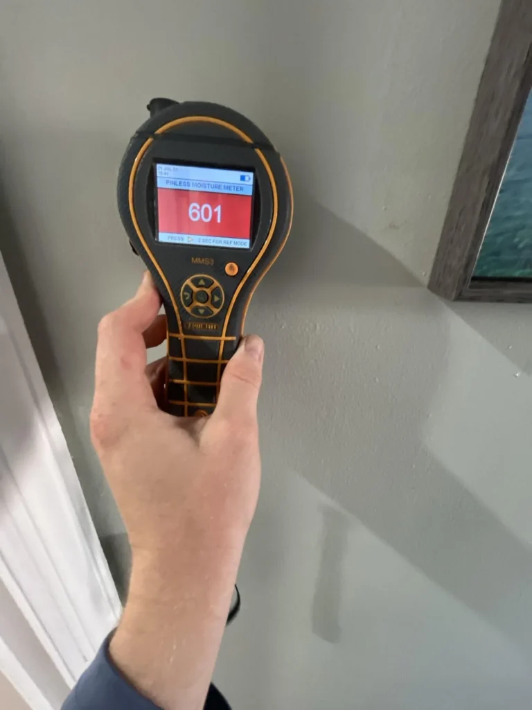 Hand holding a Protimeter MMS3 pinless moisture meter displaying a critically high reading of 601 on a red screen, held against a gray interior wall beside a framed picture