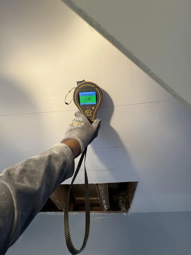 Gloved hand holding a Protimeter MMS3 pinless moisture meter displaying a reading of 136 against a ceiling wall junction above a rectangular drywall cutout exposing water-damaged floor joists