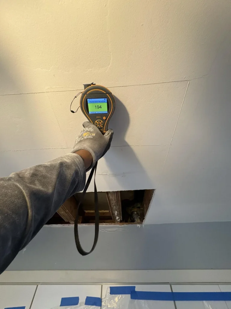 Gloved hand holding a Protimeter MMS3 pinless moisture meter reading 194 pressed against a ceiling above a rectangular access hole cut into drywall, with plastic sheeting and blue tape protecting the kitchen countertops below
