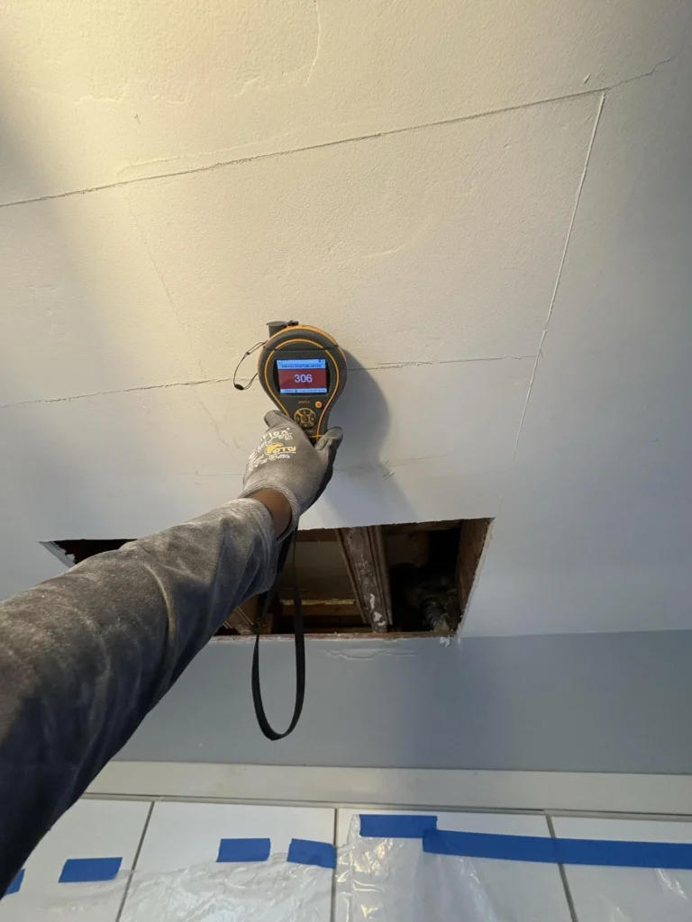 Gloved hand holding a Protimeter MMS3 pinless moisture meter showing a reading of 306 against a ceiling surface above a rectangular drywall access hole exposing water-damaged joists, with plastic sheeting protecting the kitchen cabinets below