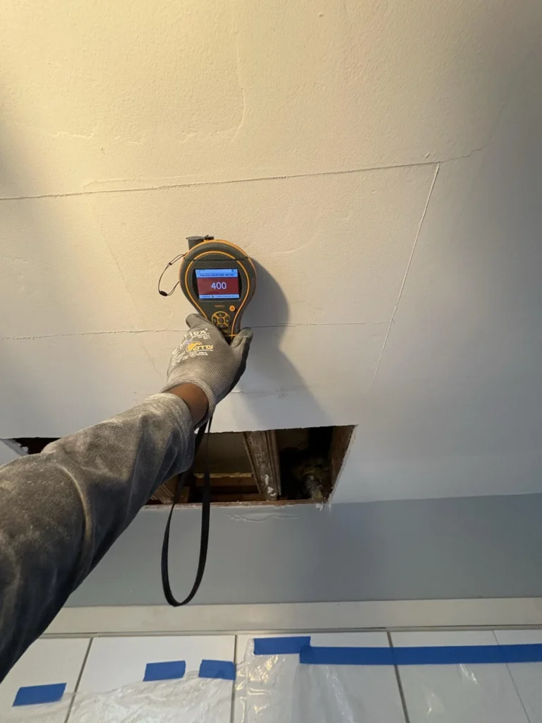 Gloved hand holding a Protimeter MMS3 pinless moisture meter displaying a maxed-out reading of 400 against a ceiling above a drywall cutout, indicating severely saturated materials, with blue tape and plastic containment sheeting visible below