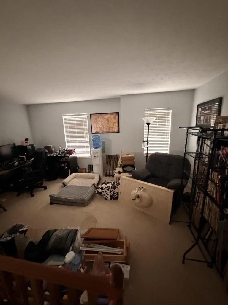 Cluttered upper-floor loft room with beige carpet, two windows with blinds, recliner armchair, water cooler, large metal shelving unit filled with vinyl records and books, desk with computer monitor, and various personal items