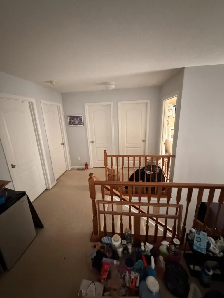 Second-floor hallway with beige carpet, three white panel doors leading to bedrooms, open door showing boxes inside, wood stair railing, smoke detector on ceiling, and cleaning supplies cluttered at the top of the stairs