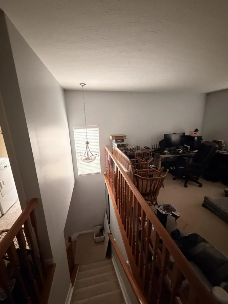 View from top of carpeted stairs looking down at a loft area with oak wood railing, pendant light fixture, washer visible to the left, and upper floor room cluttered with furniture and electronics