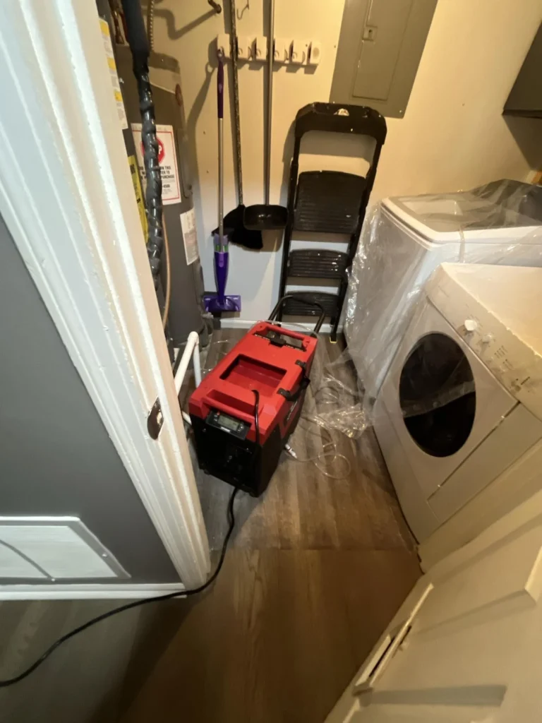 Utility room or laundry area with a red industrial dehumidifier running on the floor next to a front-load dryer still in plastic, cleaning supplies, and a water heater during water damage drying